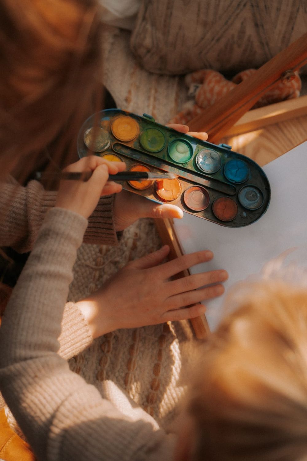 Two children painting with watercolors, sharing a colorful paint palette and brushes on white paper.