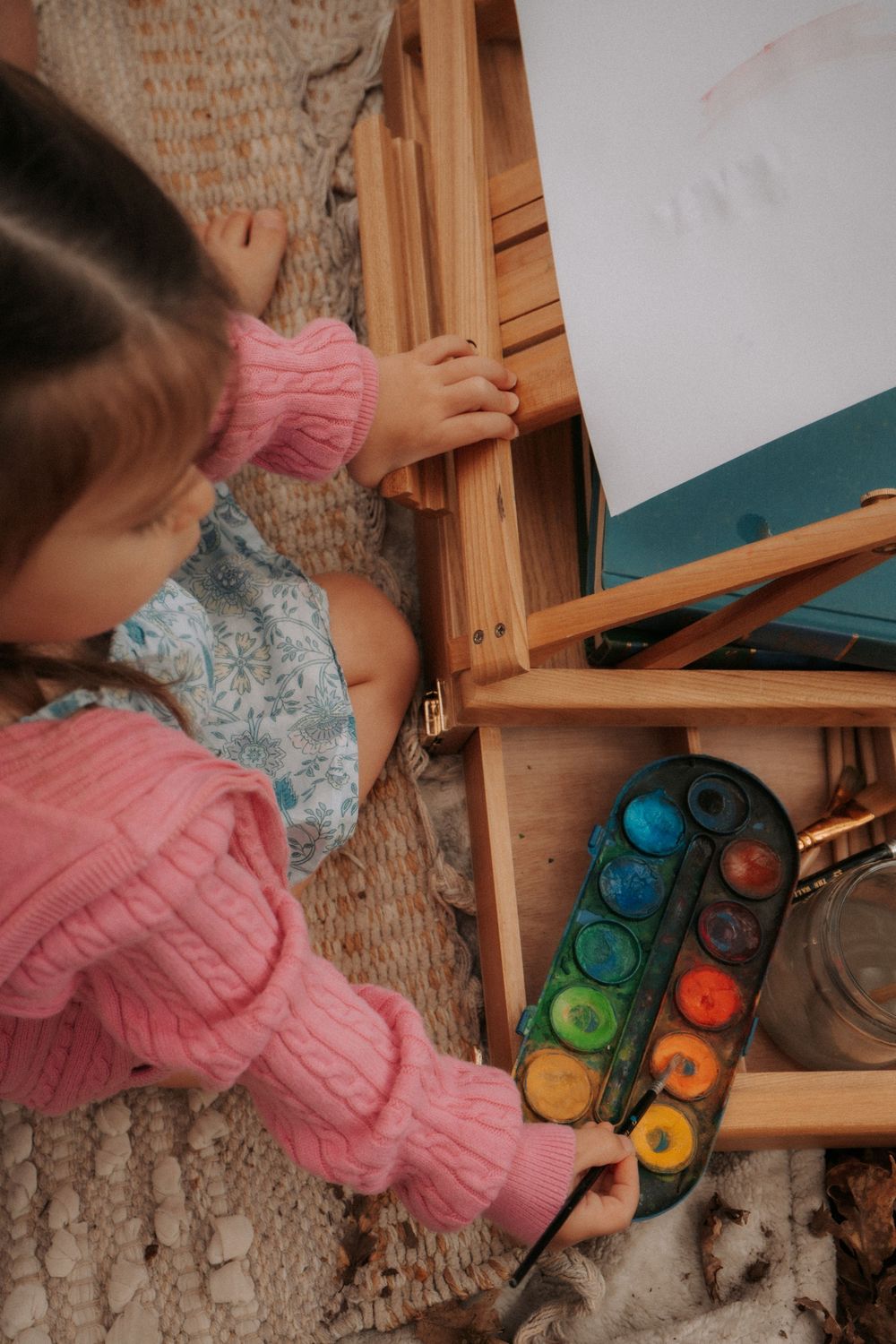 Young girl in pink sweater painting with watercolors on a wooden easel, viewed from above.