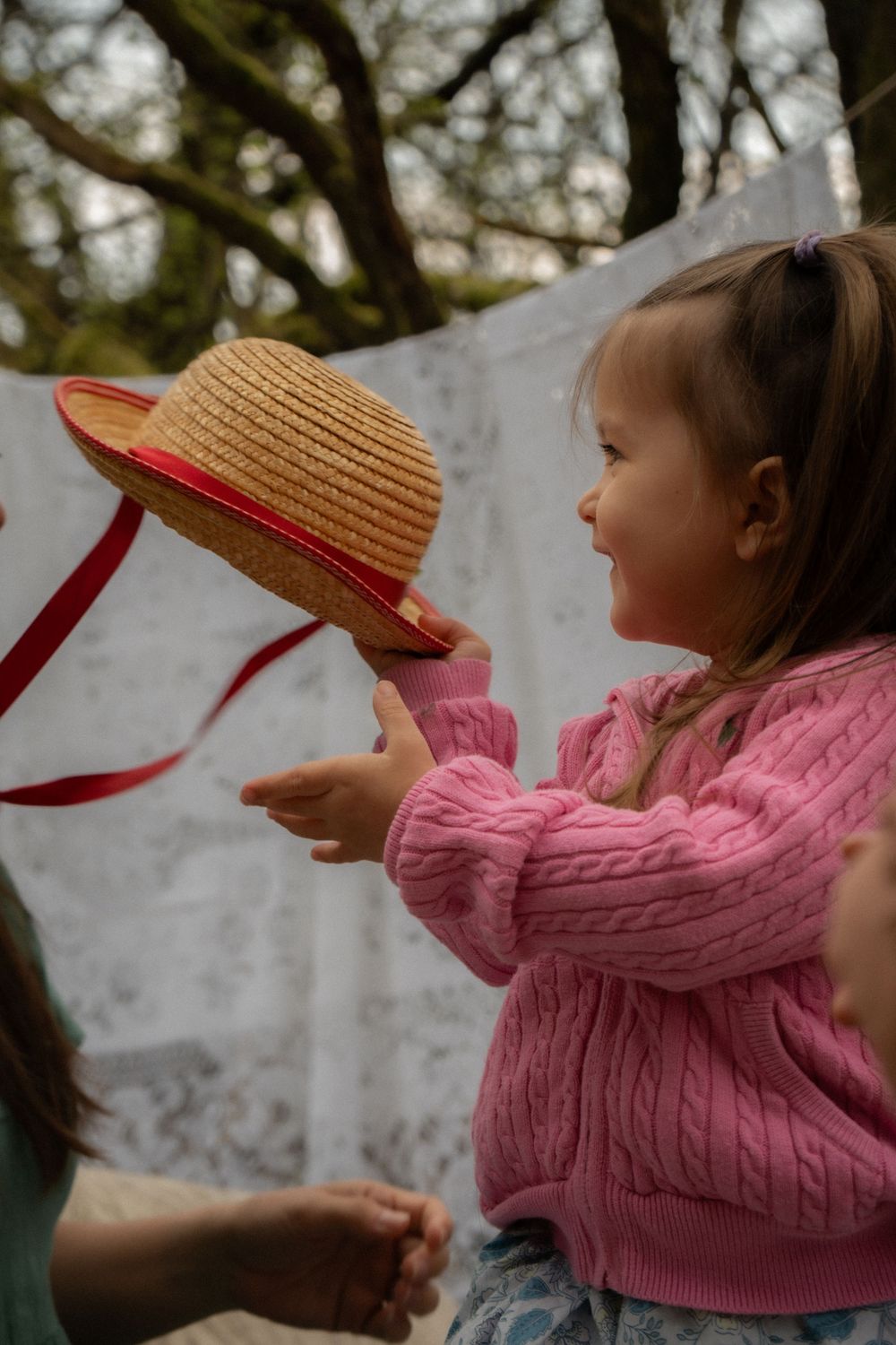 Young girl in pink sweater holding a straw hat with red ribbon outdoors near hanging white fabric.