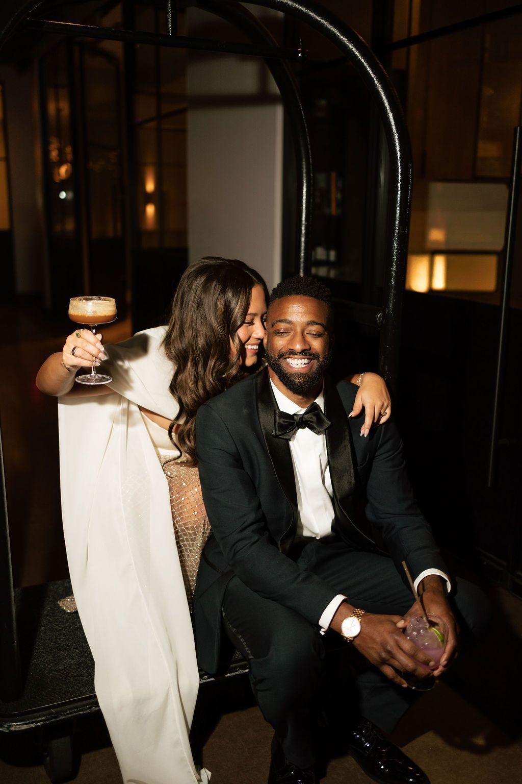 Bride in white gown and groom in black tuxedo share a joyful moment, holding cocktails at an elegant evening reception.