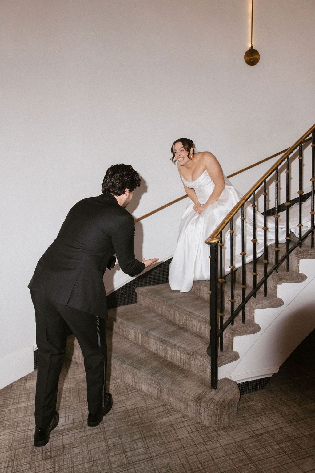 Bride in white gown descends elegant staircase toward groom in black suit holding vows at wedding venue.