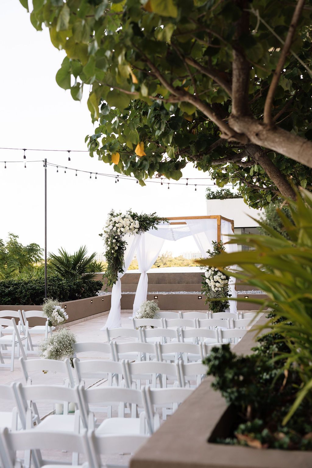 Outdoor wedding ceremony setup with white chairs, floral arch, string lights, and lush greenery under a clear sky.