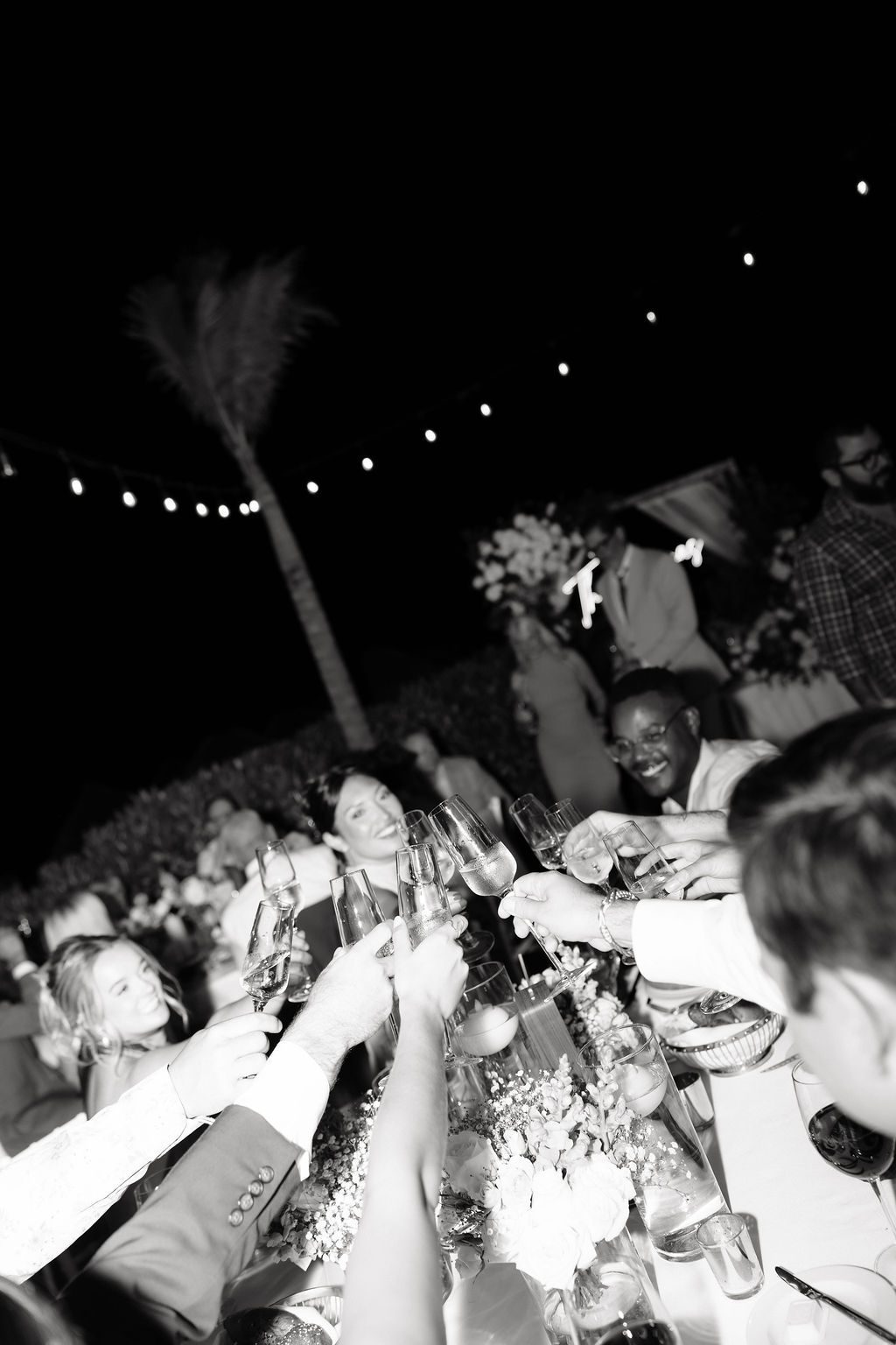 Black and white photo of wedding guests raising glasses in a toast at an outdoor evening reception with string lights.