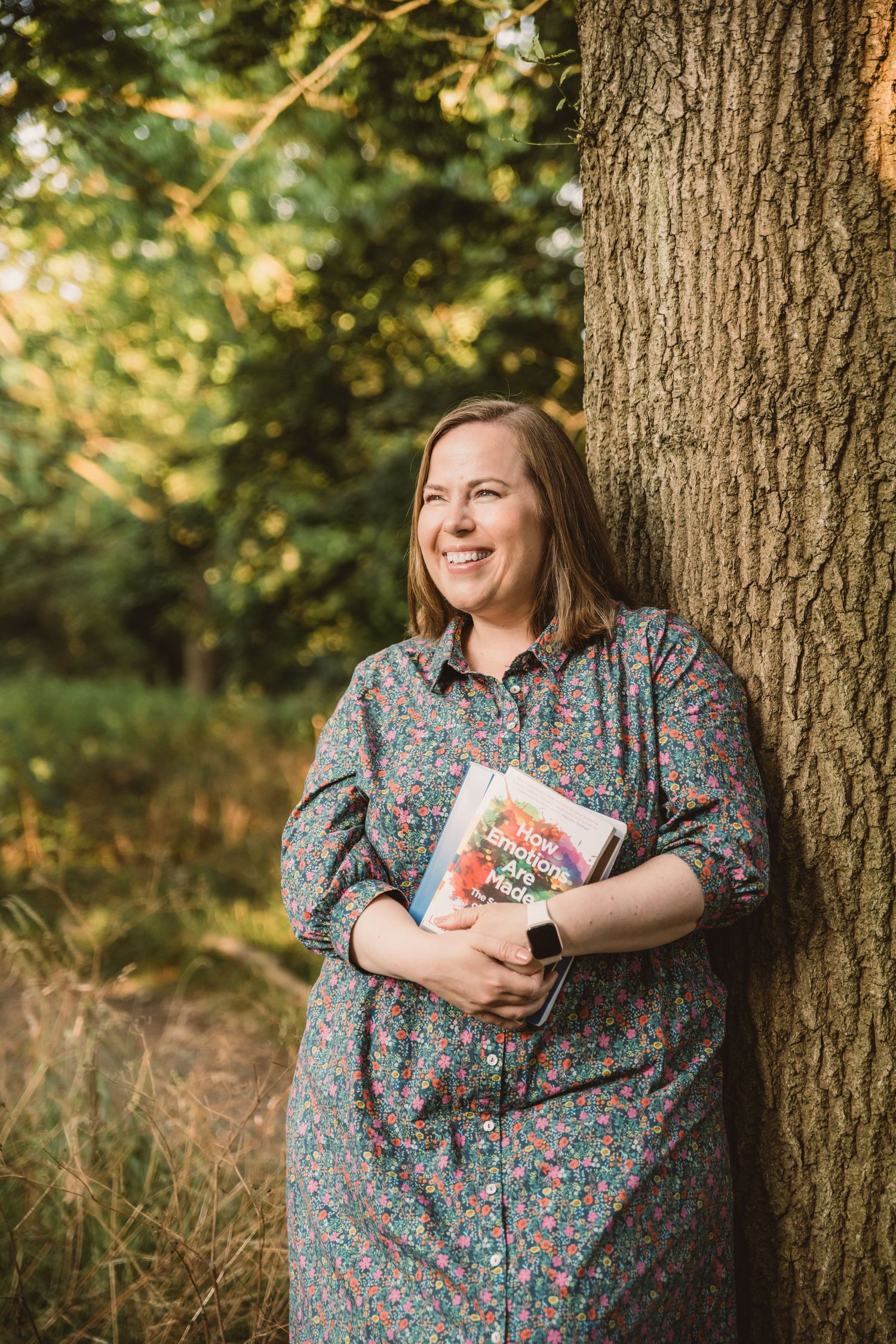 Smiling woman in floral dress holding a book, leaning against a tree in a sunlit forest setting.