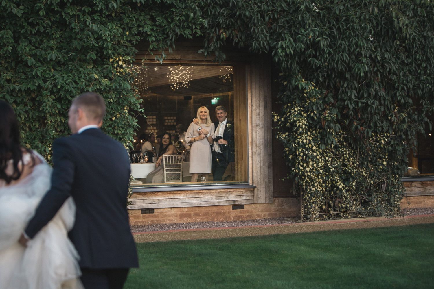 Wedding couple walks toward ivy-covered venue entrance where guests gather in doorway on lush green lawn.