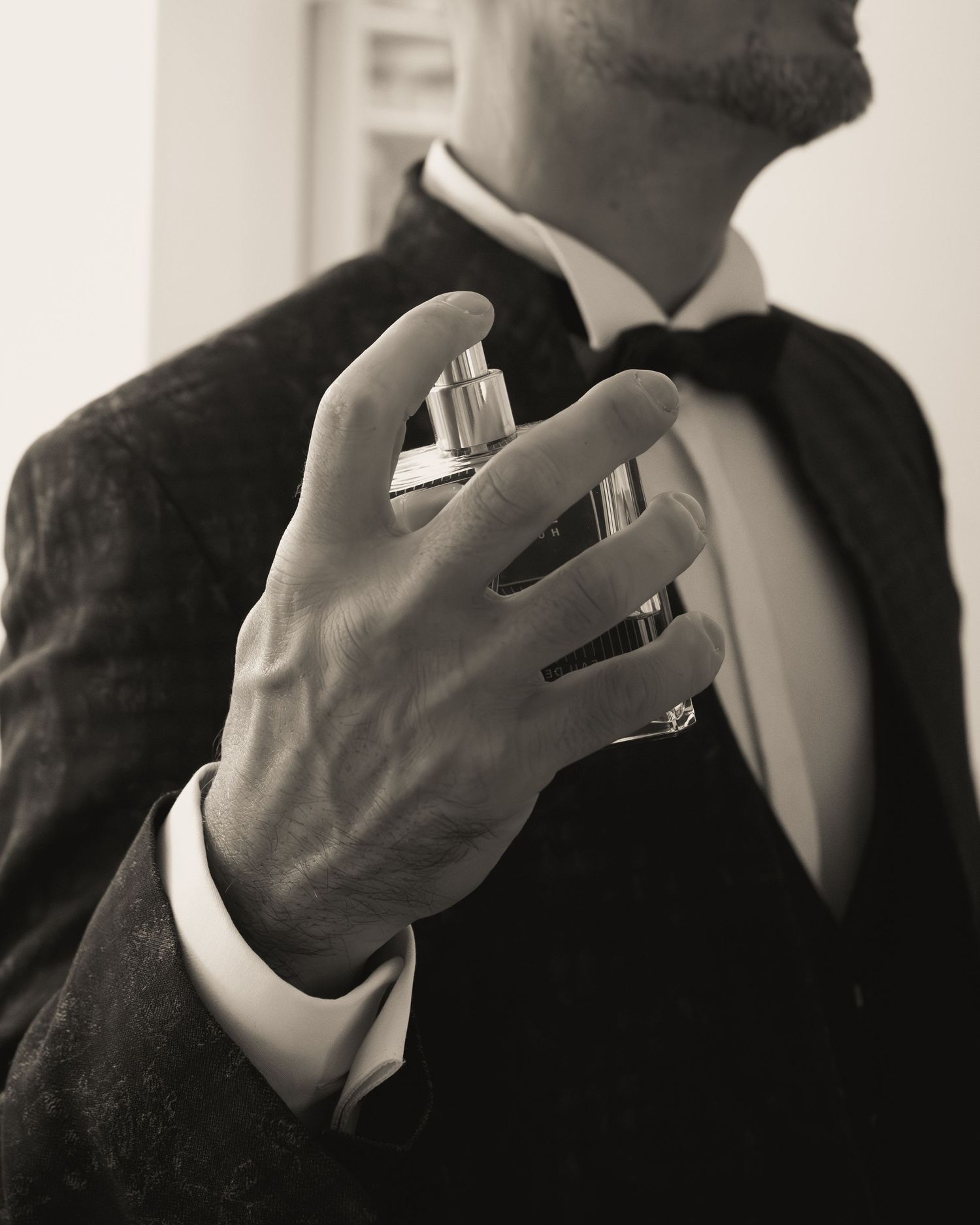 Black and white close-up of groom in velvet tuxedo applying cologne before wedding ceremony.