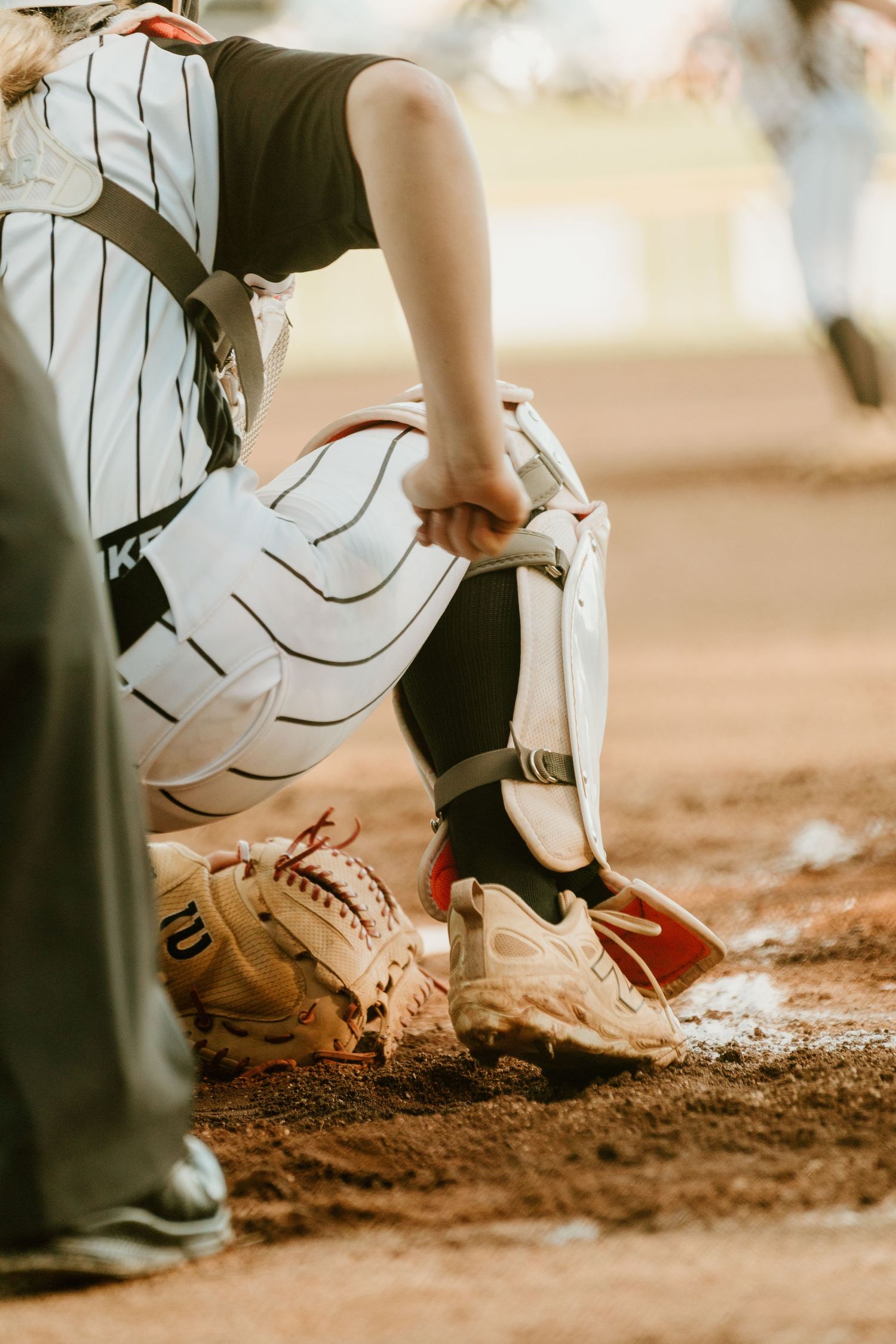 Baseball catcher in full gear kneeling at home plate with Wilson glove on dirt field during game.