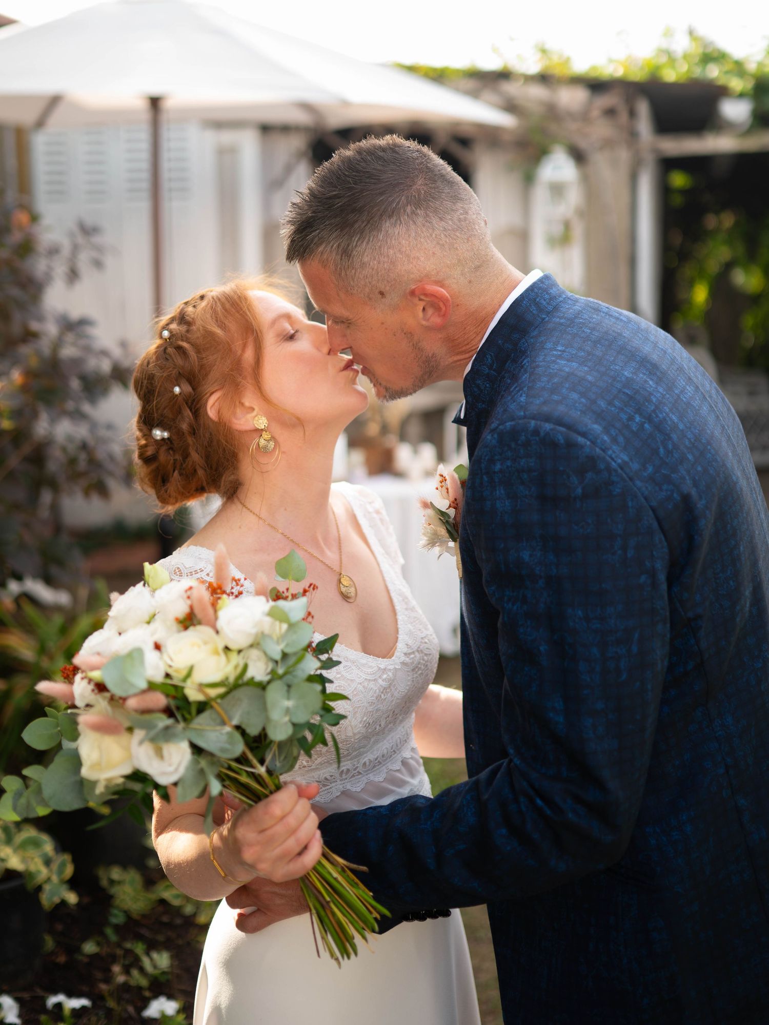 Les mariés s'embrassent tendrement en plein air ; la mariée tient un bouquet de fleurs blanches et roses.