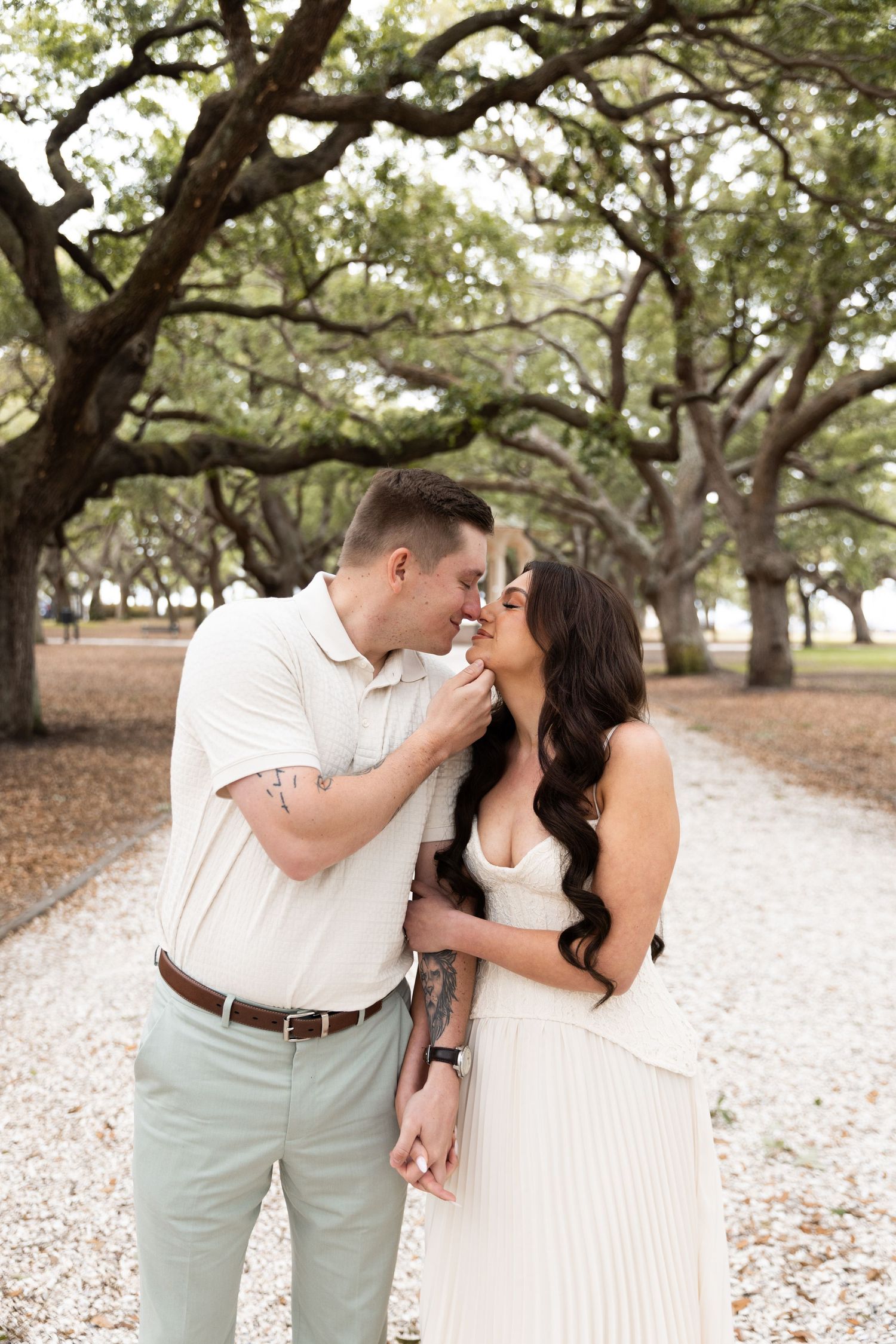 Couple sharing a romantic moment on a tree-lined path, man in white shirt and woman in white dress.