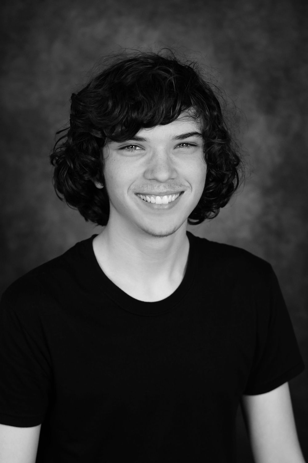 Black and white portrait of a smiling young man with curly dark hair wearing a black t-shirt.