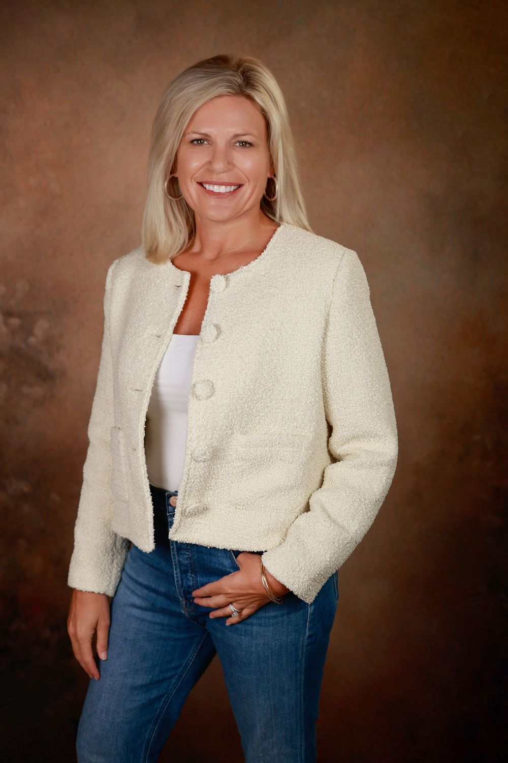 Smiling blonde woman in a cream blazer and jeans posing confidently against a warm brown studio background.