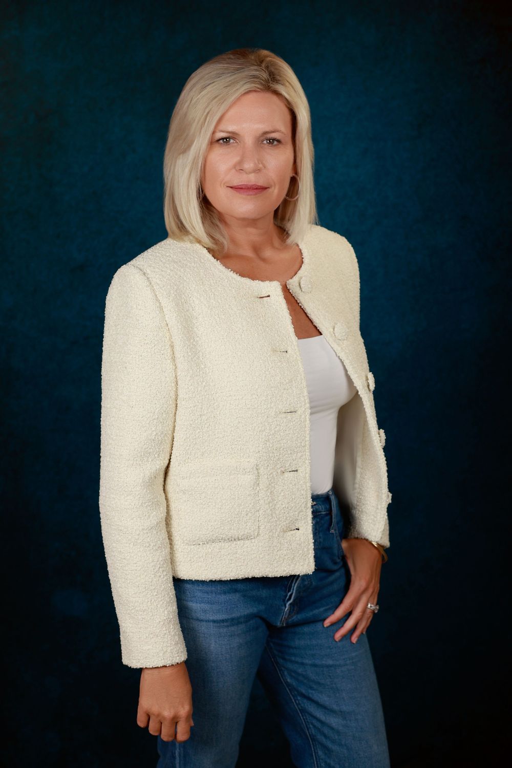 Professional portrait of a blonde woman in a cream blazer and jeans against a blue background.