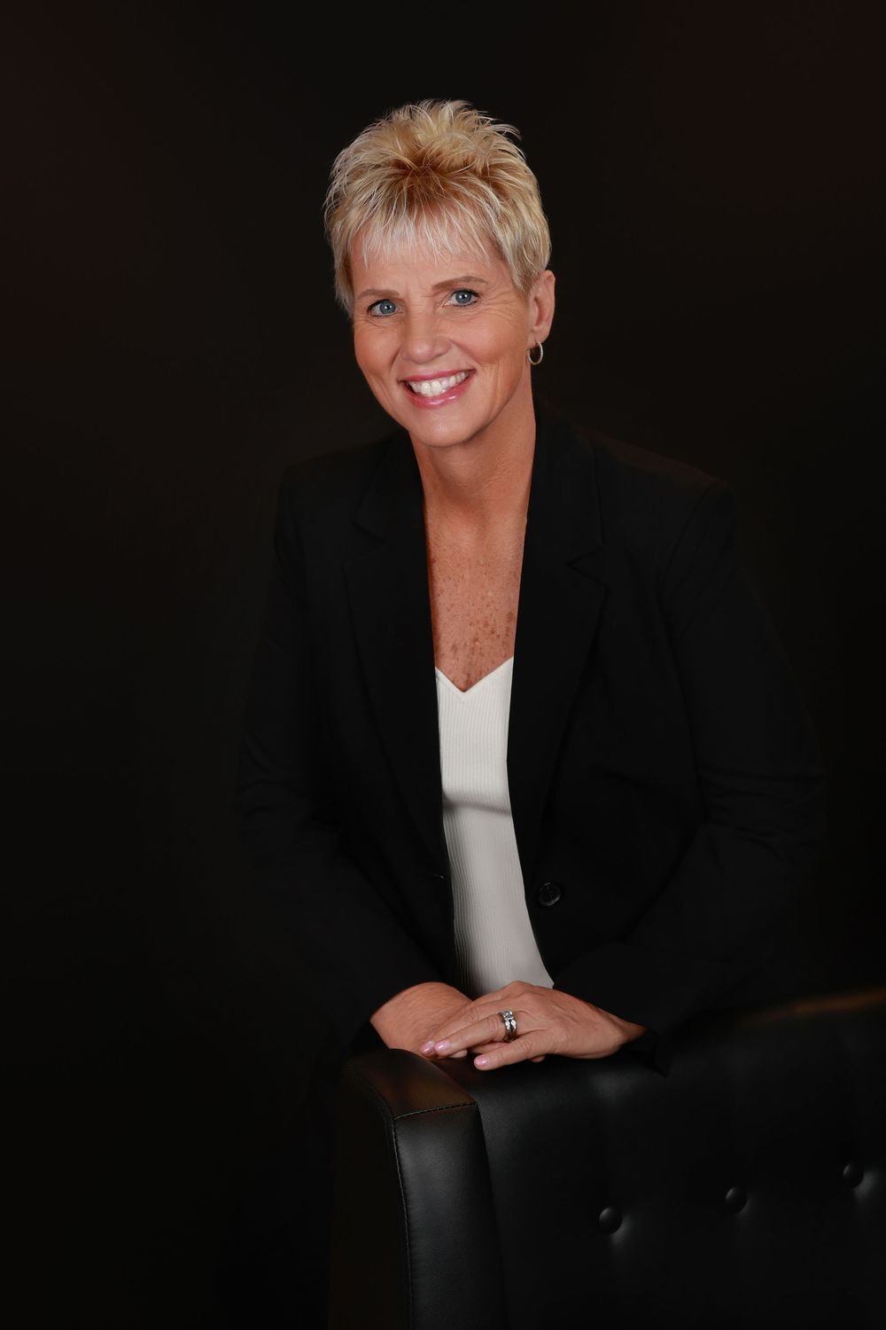 Professional blonde woman in a black blazer smiling warmly while leaning on a chair against a dark background.