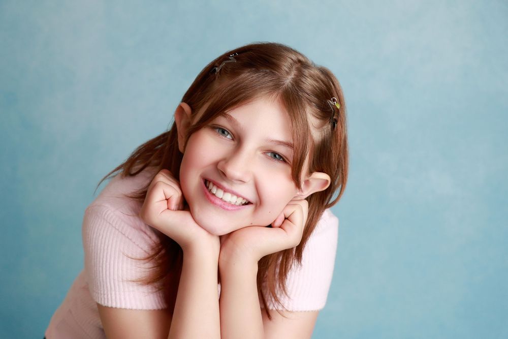 Young girl with brown hair smiling with hands on cheeks against a light blue background.