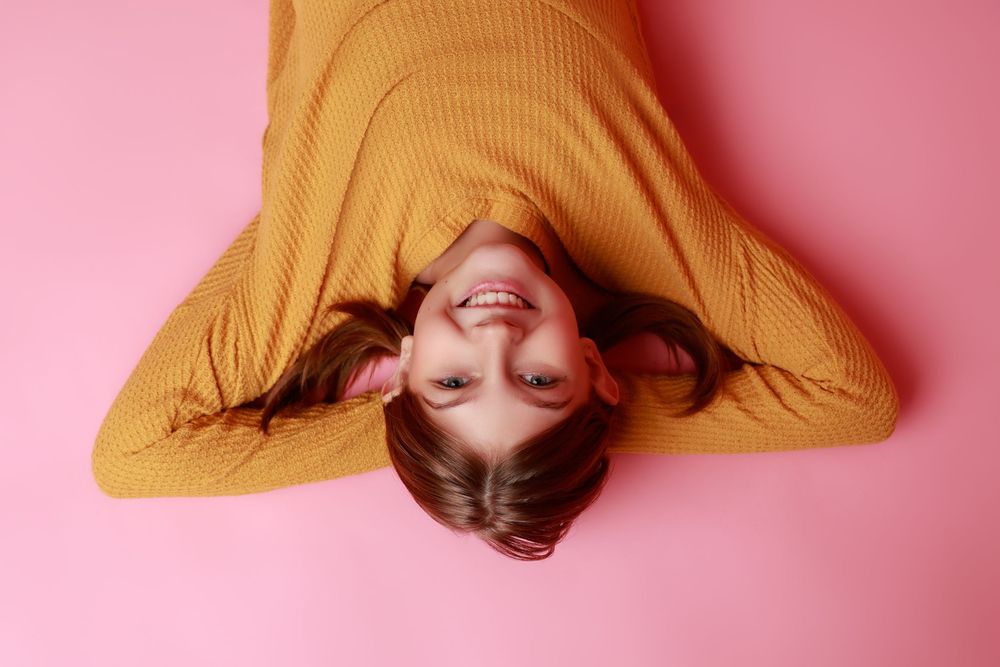 Happy young woman in a mustard yellow sweater lying on a pink background, smiling upside down at the camera.