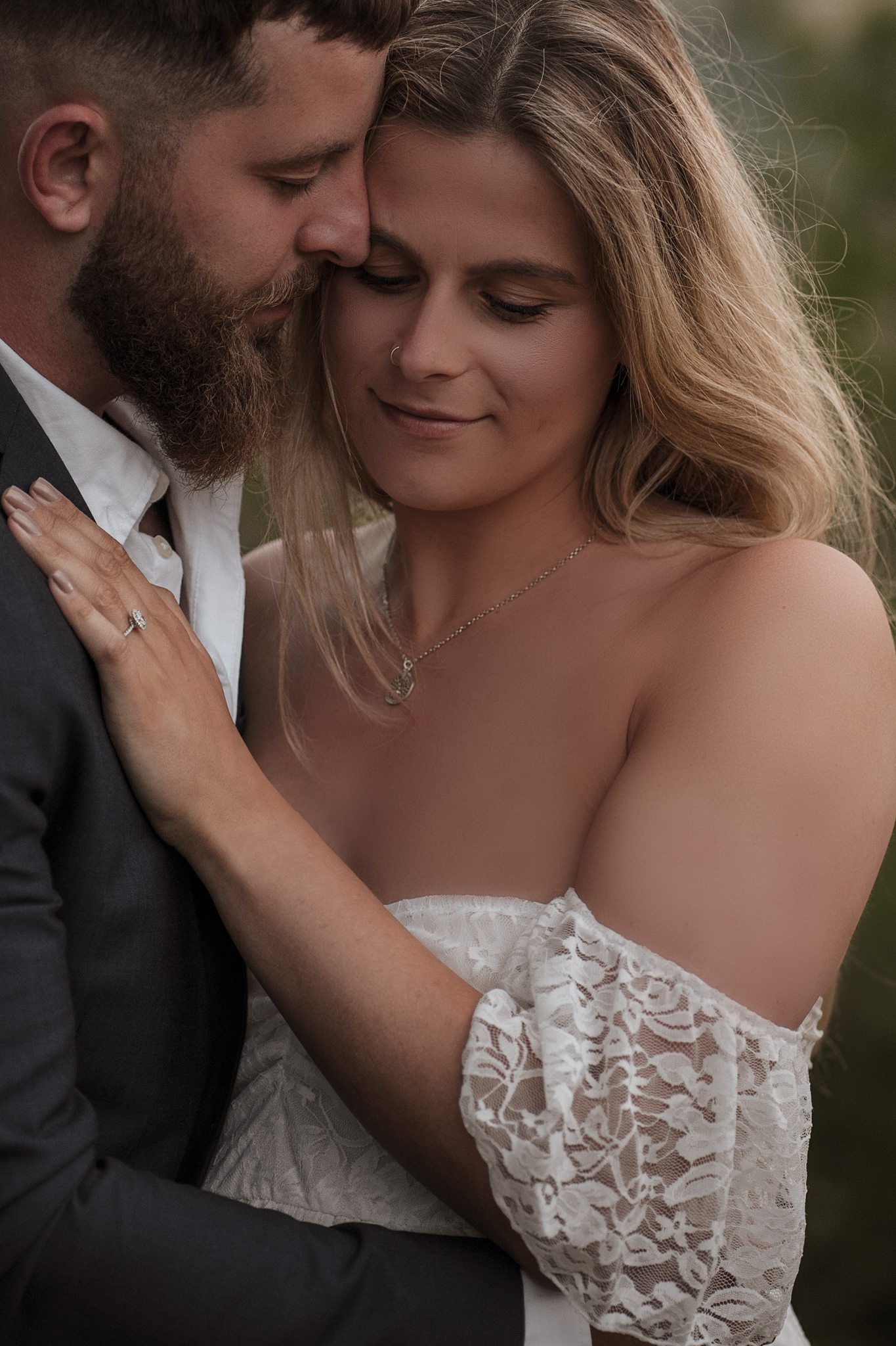A bride in lace off-shoulder gown embraces bearded groom in dark suit, sharing an intimate romantic moment.