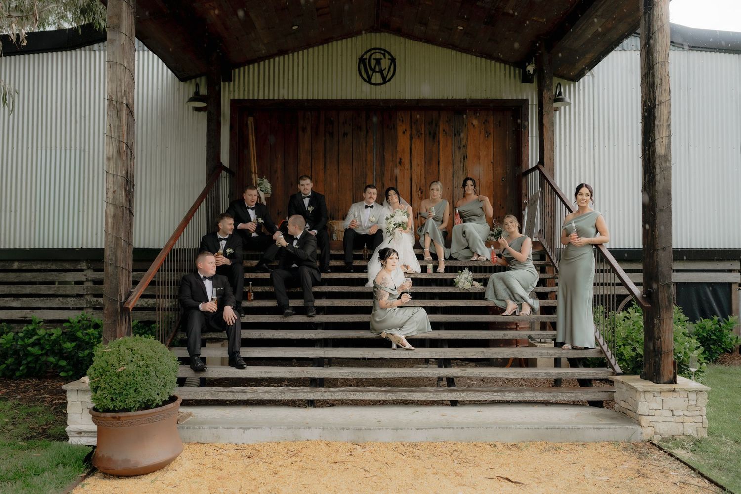 Wedding party posed on rustic barn steps, bride and groom surrounded by groomsmen and bridesmaids in sage green gowns.