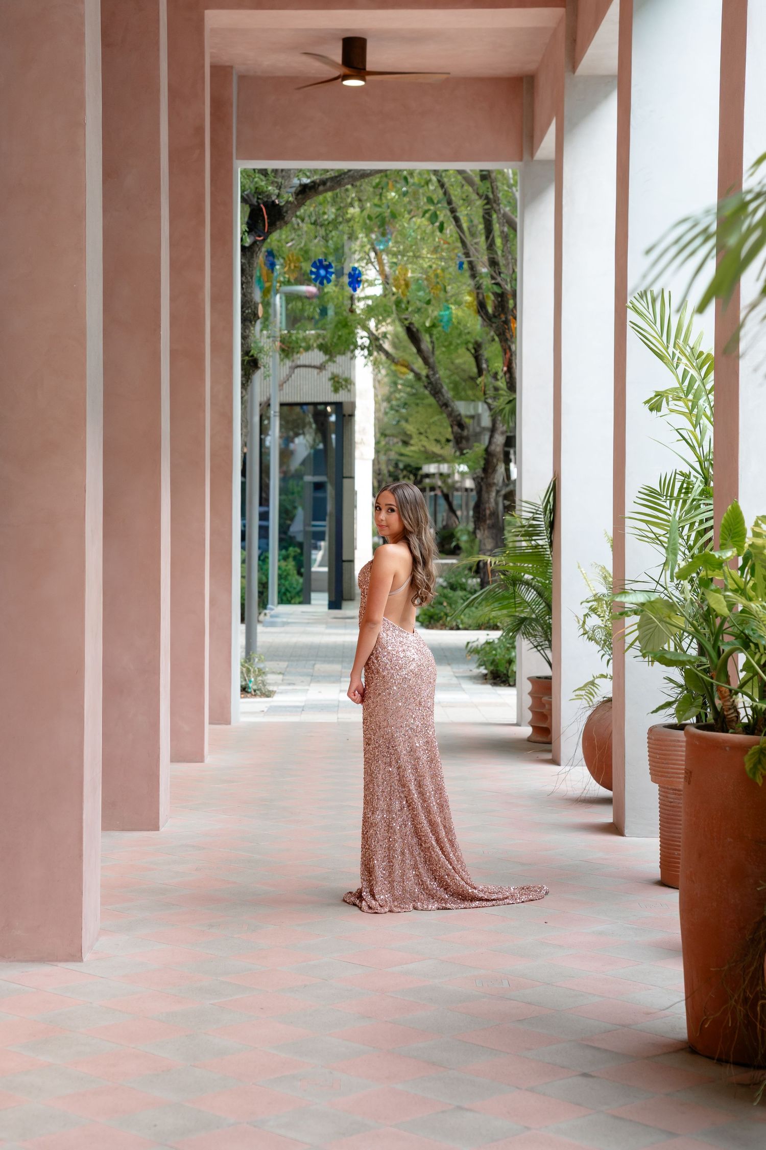 Woman in elegant rose gold sequin gown posing in a pink columned corridor with lush tropical greenery.