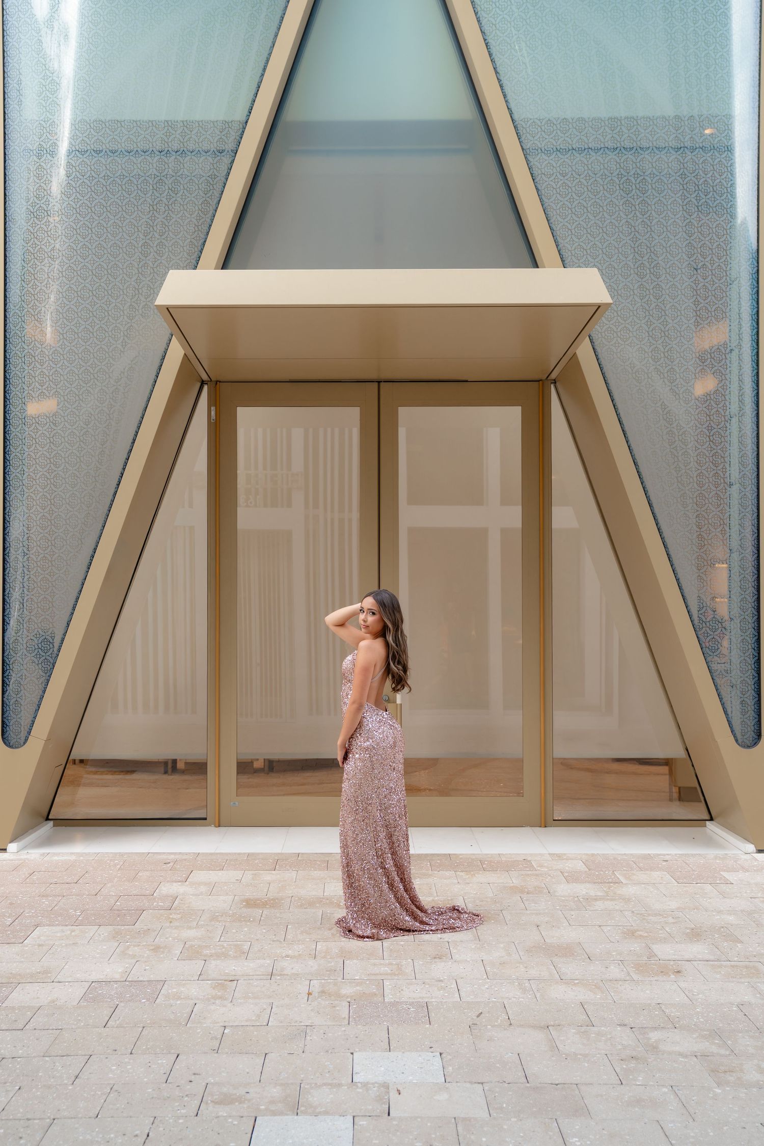 Woman in elegant pink sequin gown poses before modern architectural doorway with triangular frame on stone patio.