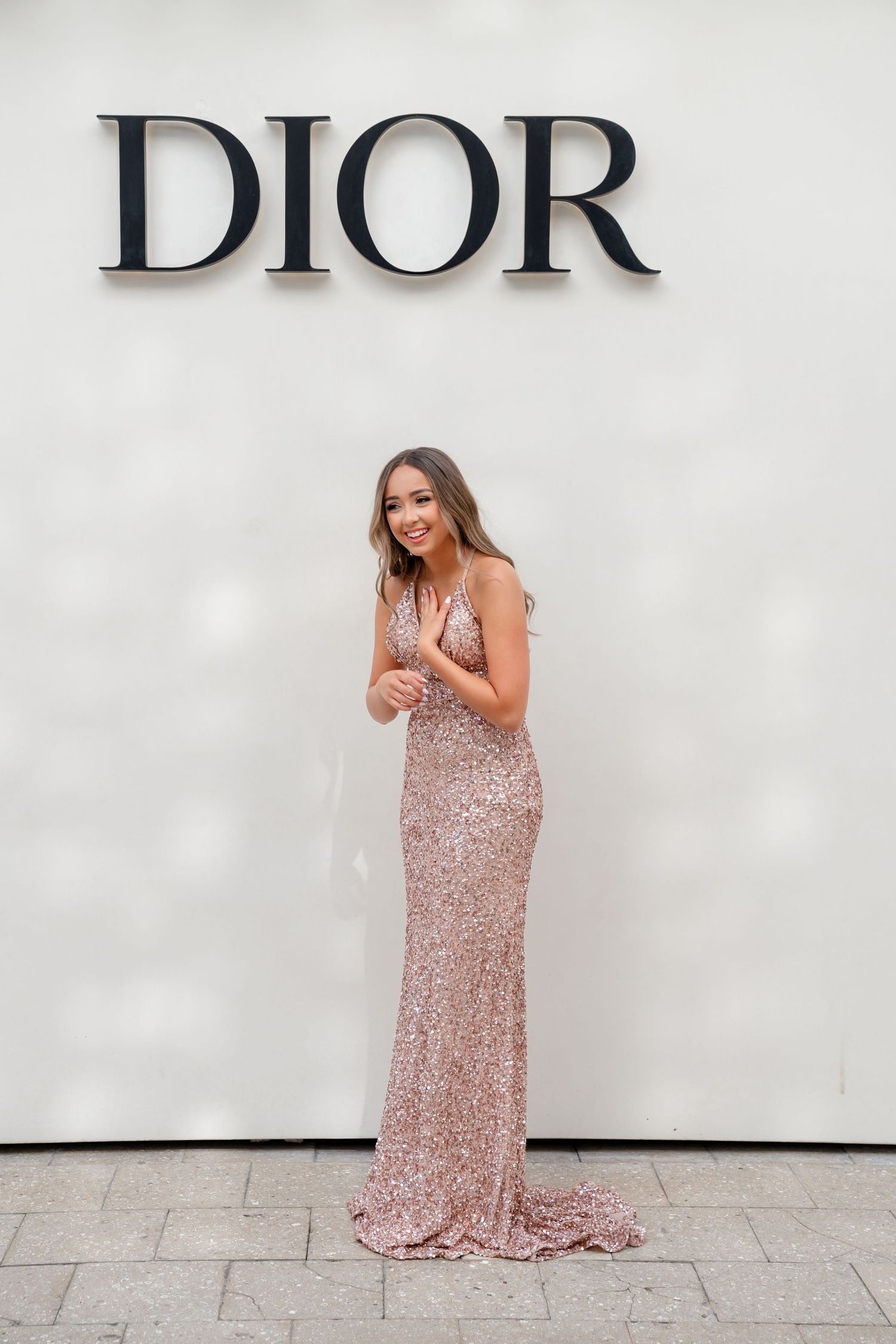 Woman in rose gold sequin gown posing in front of Dior logo at luxury fashion event.