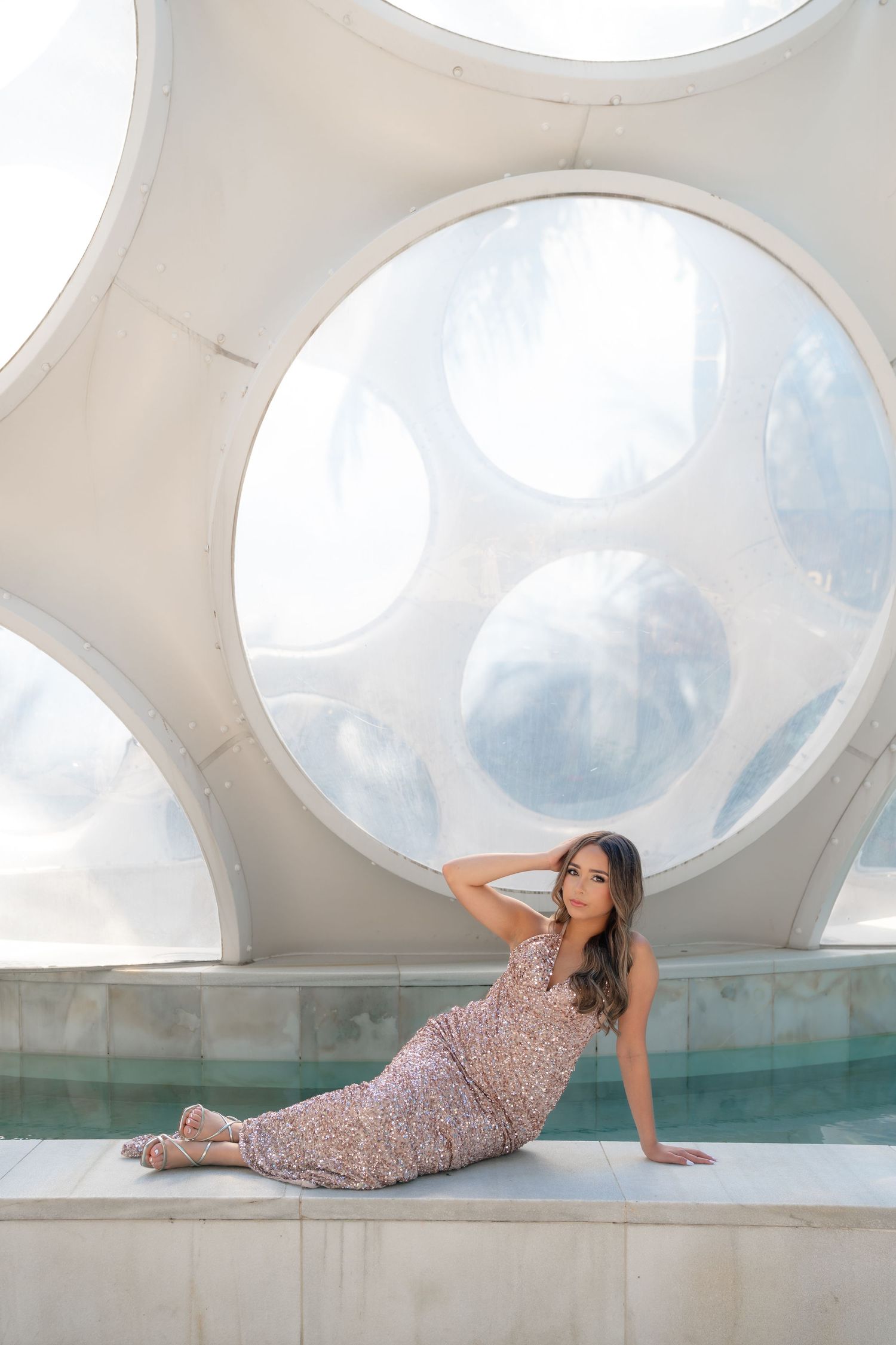 Woman in floral dress poses elegantly by a pool with circular geometric white architecture backdrop.