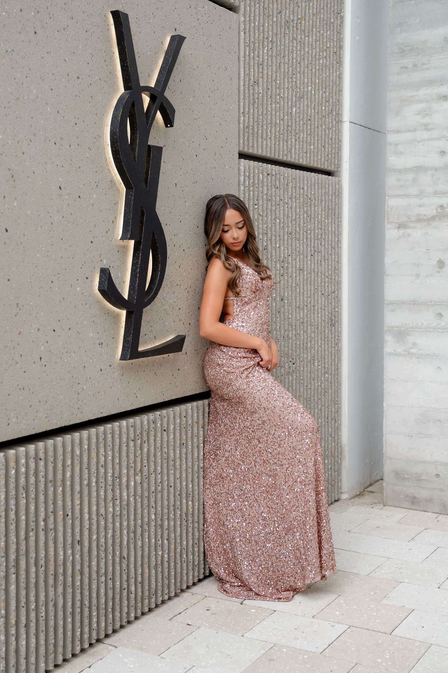 Woman in rose gold sequin gown posing beside YSL luxury brand sign on textured concrete wall.