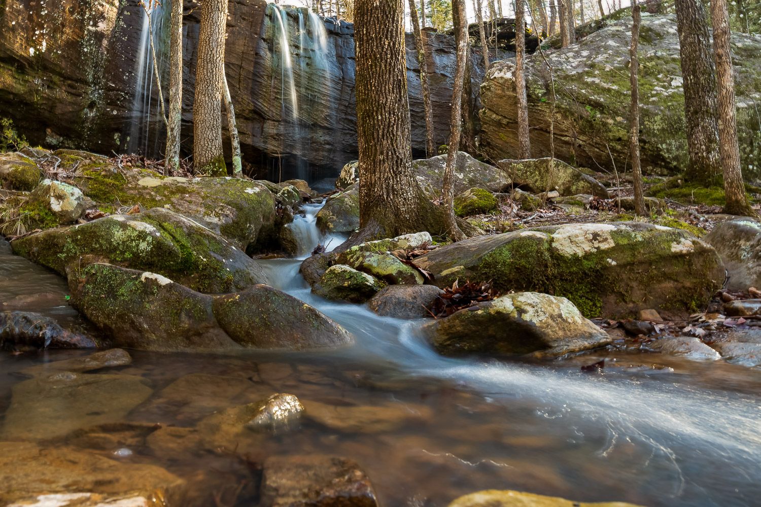 A small waterfall flows over mossy rocks through a sunlit forest stream with long exposure silky water effect. Sweden Creek area, NW AR