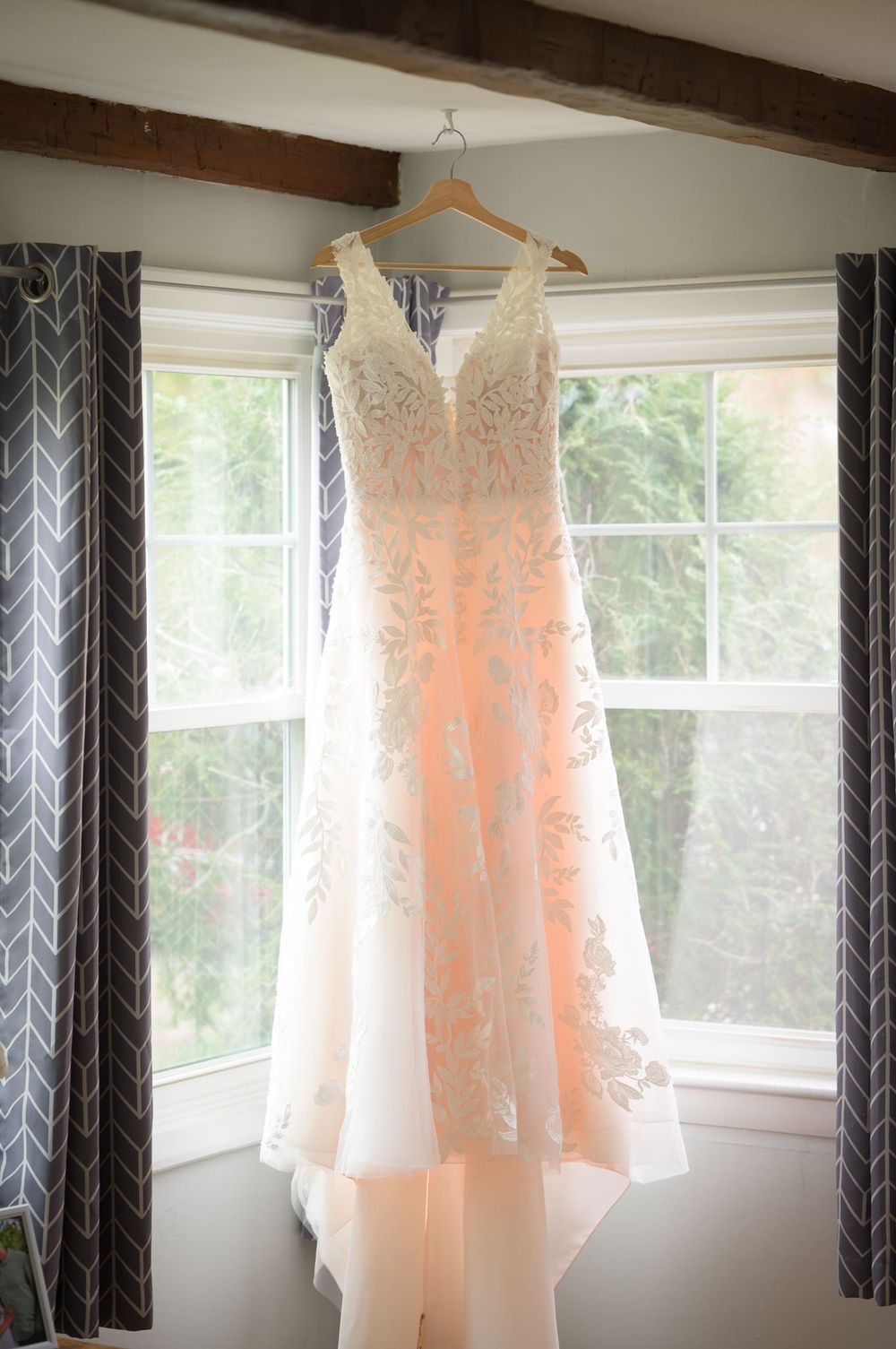 Elegant white lace wedding gown hanging on a wooden hanger in a sunlit window.