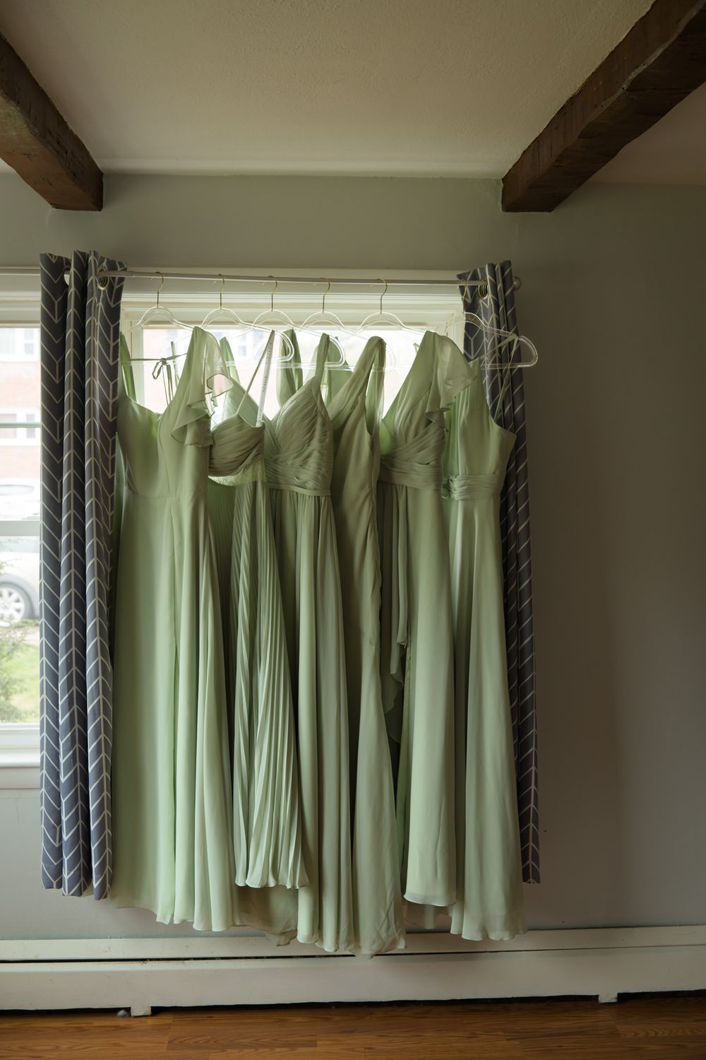 Six sage green bridesmaid dresses hanging on white hangers in front of a window with gray chevron curtains.