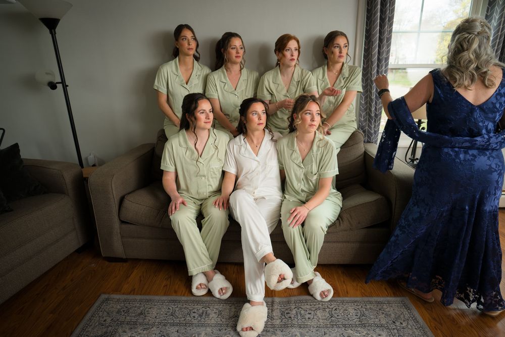 Seven bridesmaids in sage green pajamas pose with bride in white on a couch during wedding prep.