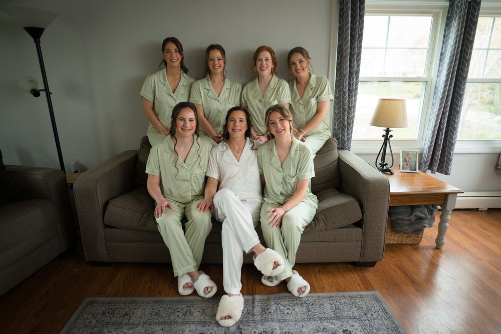 Bride in white pajamas poses with six bridesmaids in matching green sets on a gray couch indoors.