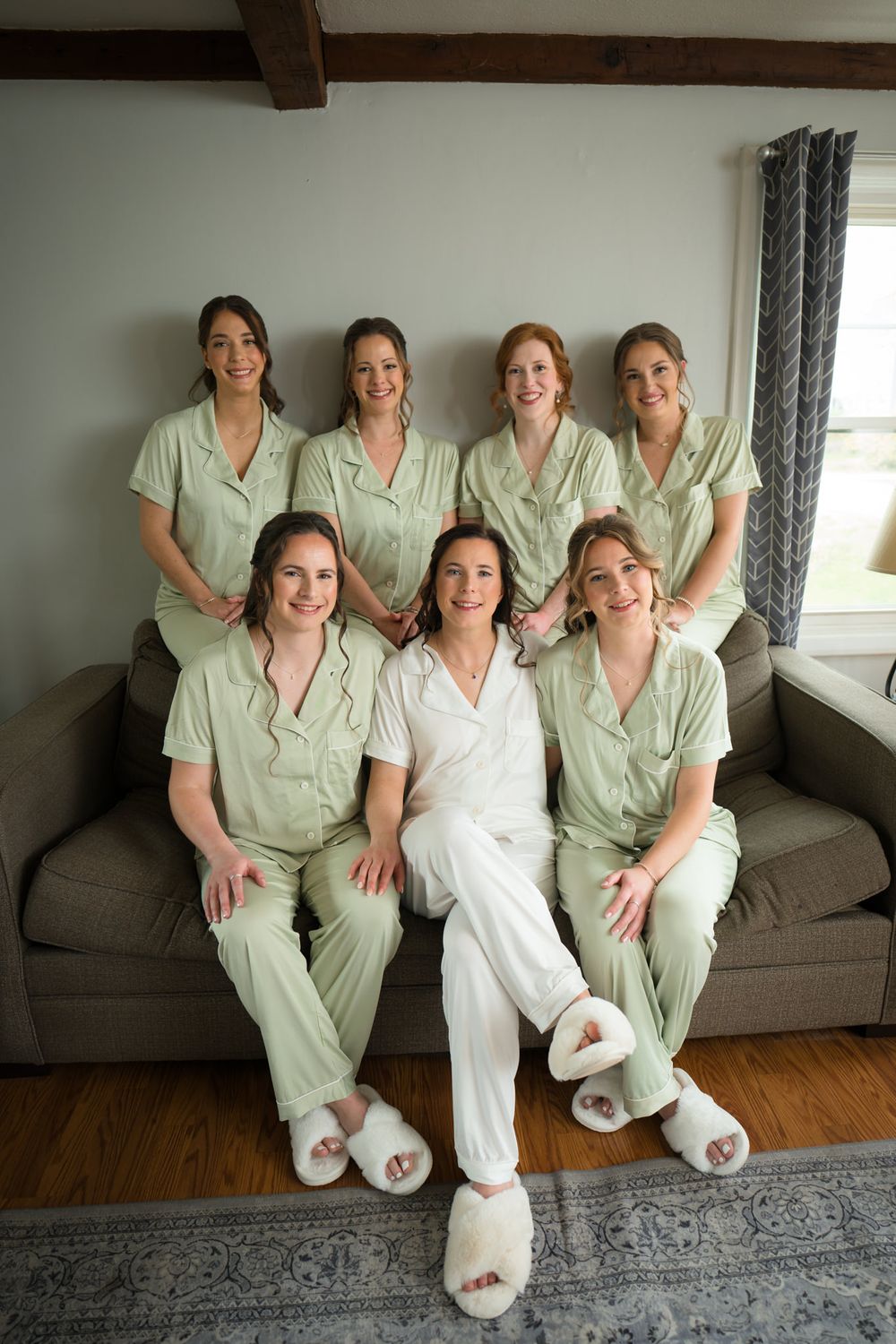 A bride in white pajamas poses with six bridesmaids in matching sage green sets on a cozy couch.