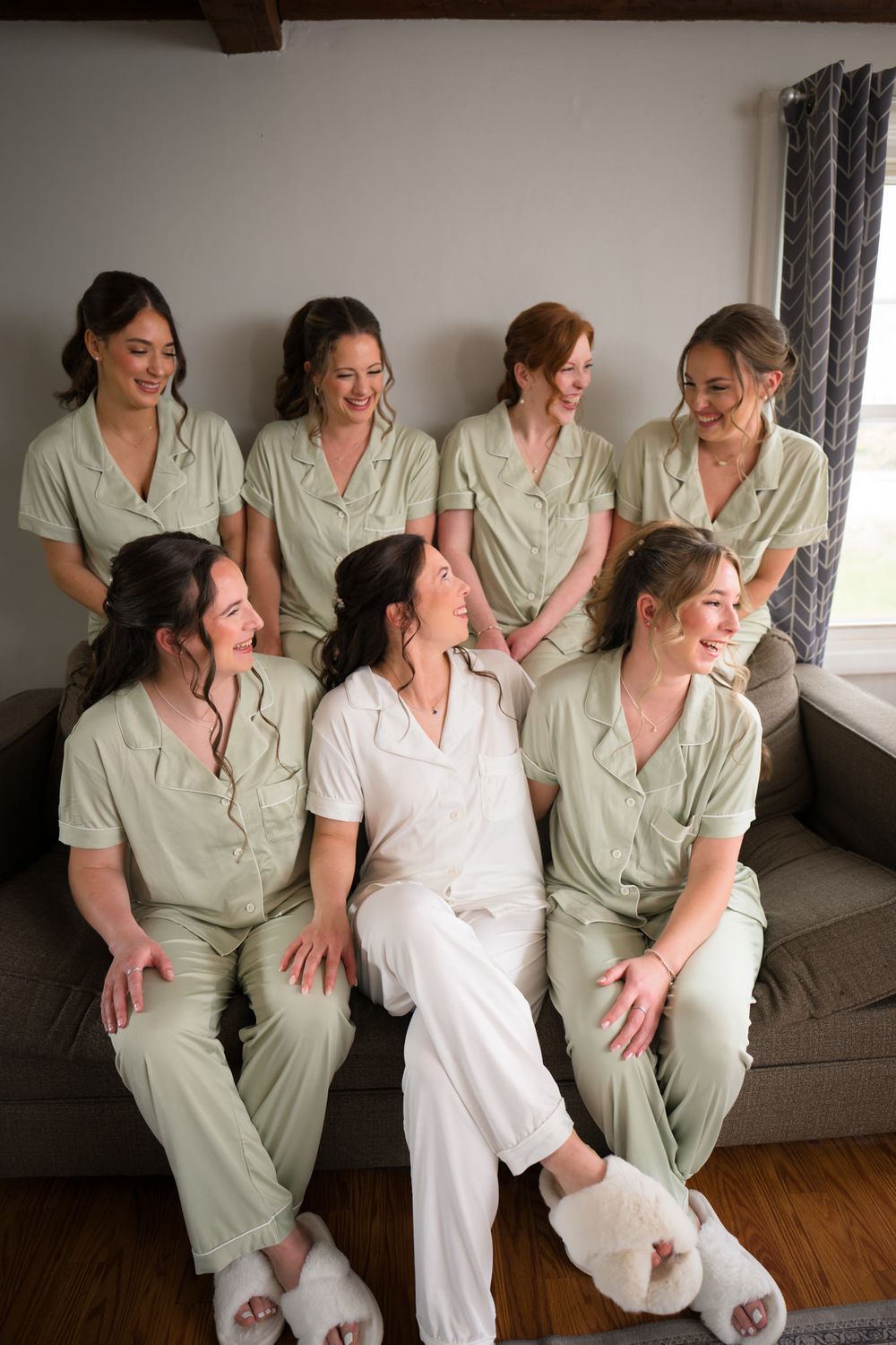 Bride in white pajamas sits with seven bridesmaids in matching sage green sets on a couch, smiling together.