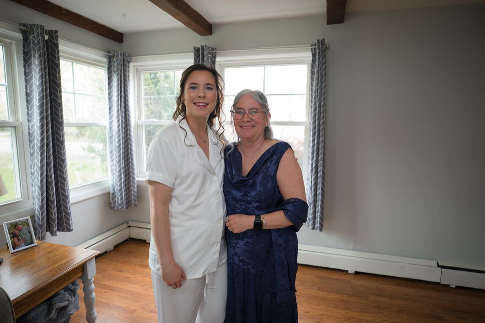 Bride in white outfit poses with older woman in navy blue dress in a bright windowed room.