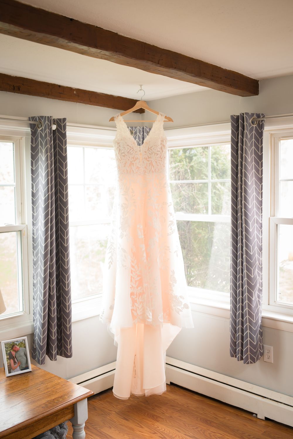 A blush pink lace wedding dress hanging on a wooden beam between two windows with gray chevron curtains.