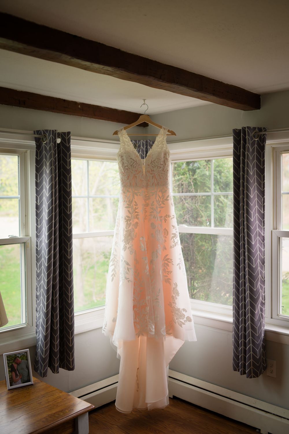 Elegant blush lace wedding dress hanging on a wooden hanger in a window with gray curtains and exposed wooden beams.
