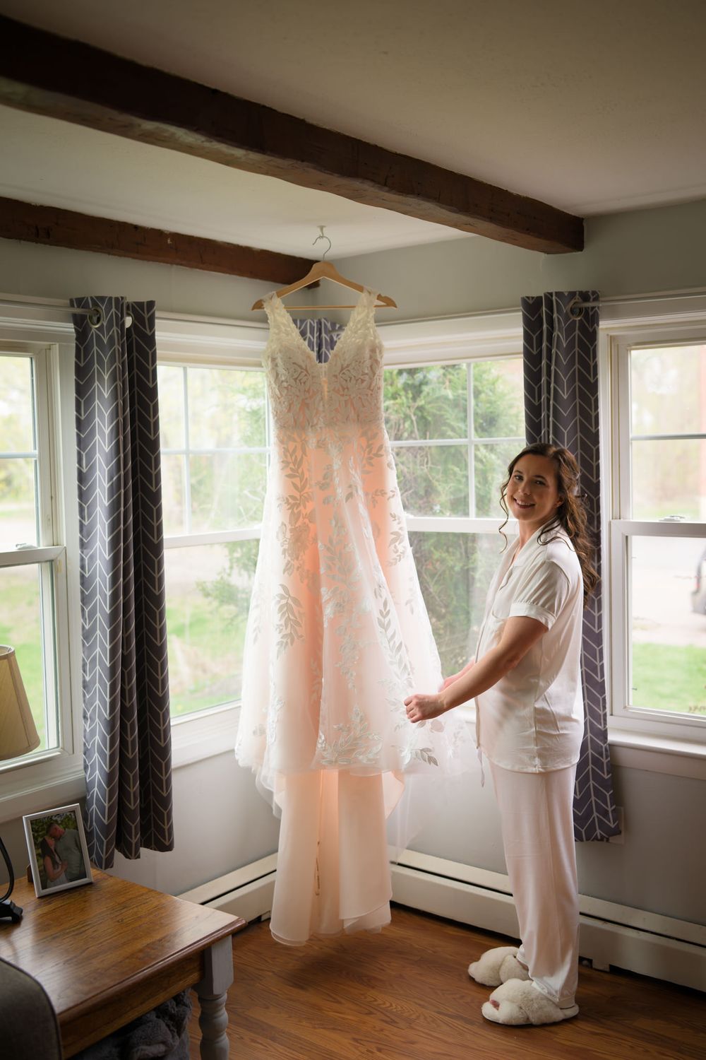 Smiling bride in white pajamas admiring her blush pink lace wedding gown hanging near sunlit windows.