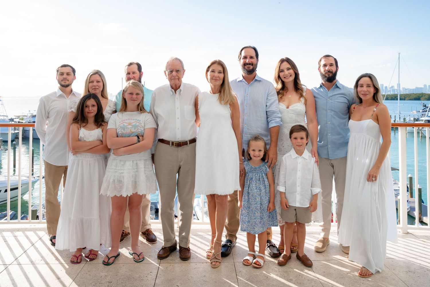 Large family group portrait on a waterfront deck, dressed in white and light blue outfits, smiling together.