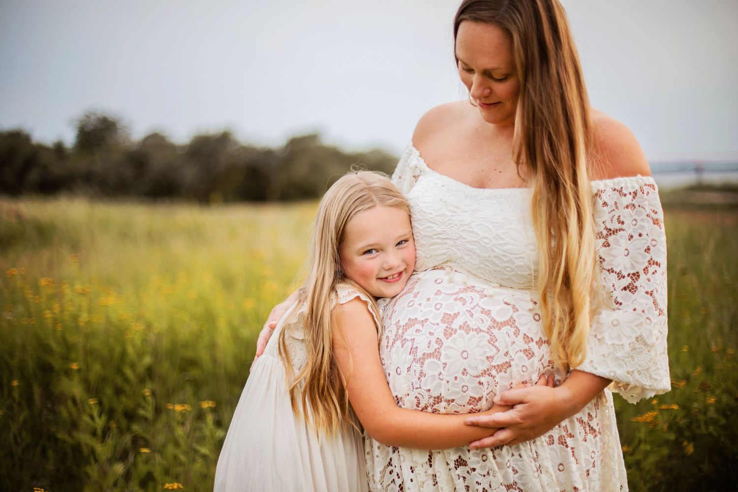 En ung flicka kramar sin gravida mammas mage i ett soligt blomsterfält, båda klädda i vita spetsklänningar.