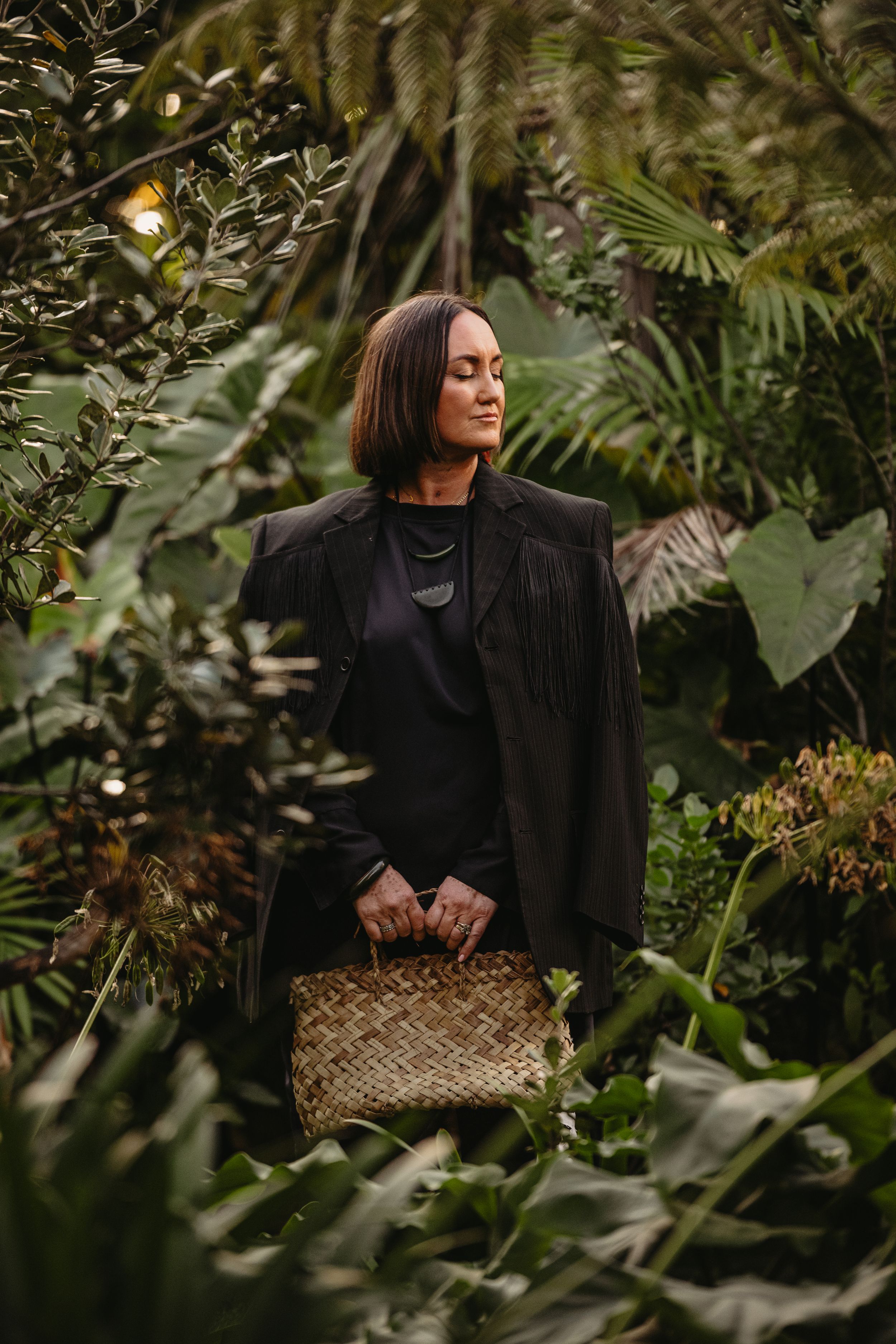Woman in black blazer holding woven basket surrounded by lush tropical greenery.