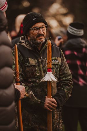 Man in camouflage jacket and black beanie holds decorated wooden staff at outdoor gathering.