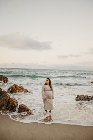Pregnant woman in beige dress standing on sandy beach with rocky shore and ocean waves at sunset.