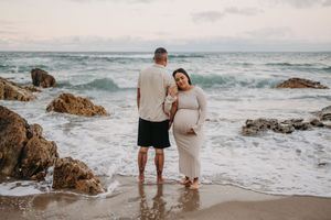 Couple standing on rocky beach with ocean waves, pregnant woman in flowing white dress cradling baby bump at sunset.