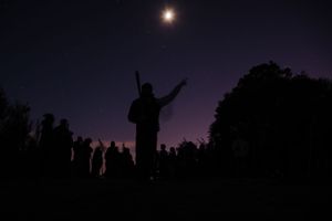 Silhouette of a person with raised arms under a full moon at twilight, surrounded by a crowd against a purple night sky.