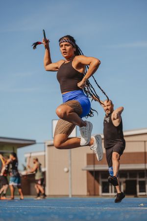 Athletic woman with dreadlocks leaping mid-air holding baton during outdoor track relay race on blue track.