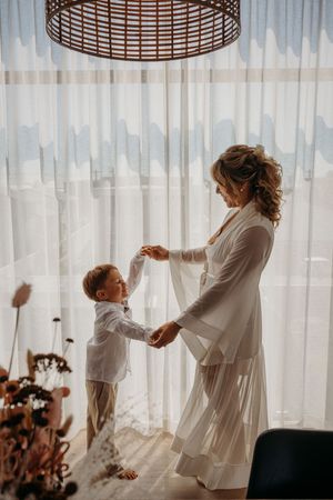 A bride in a white robe dances with a young boy in white attire near sheer curtains in a sunlit room.