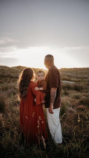 Family of three standing in golden field at sunset, parents holding baby in matching rust outfits, backlit warm glow.
