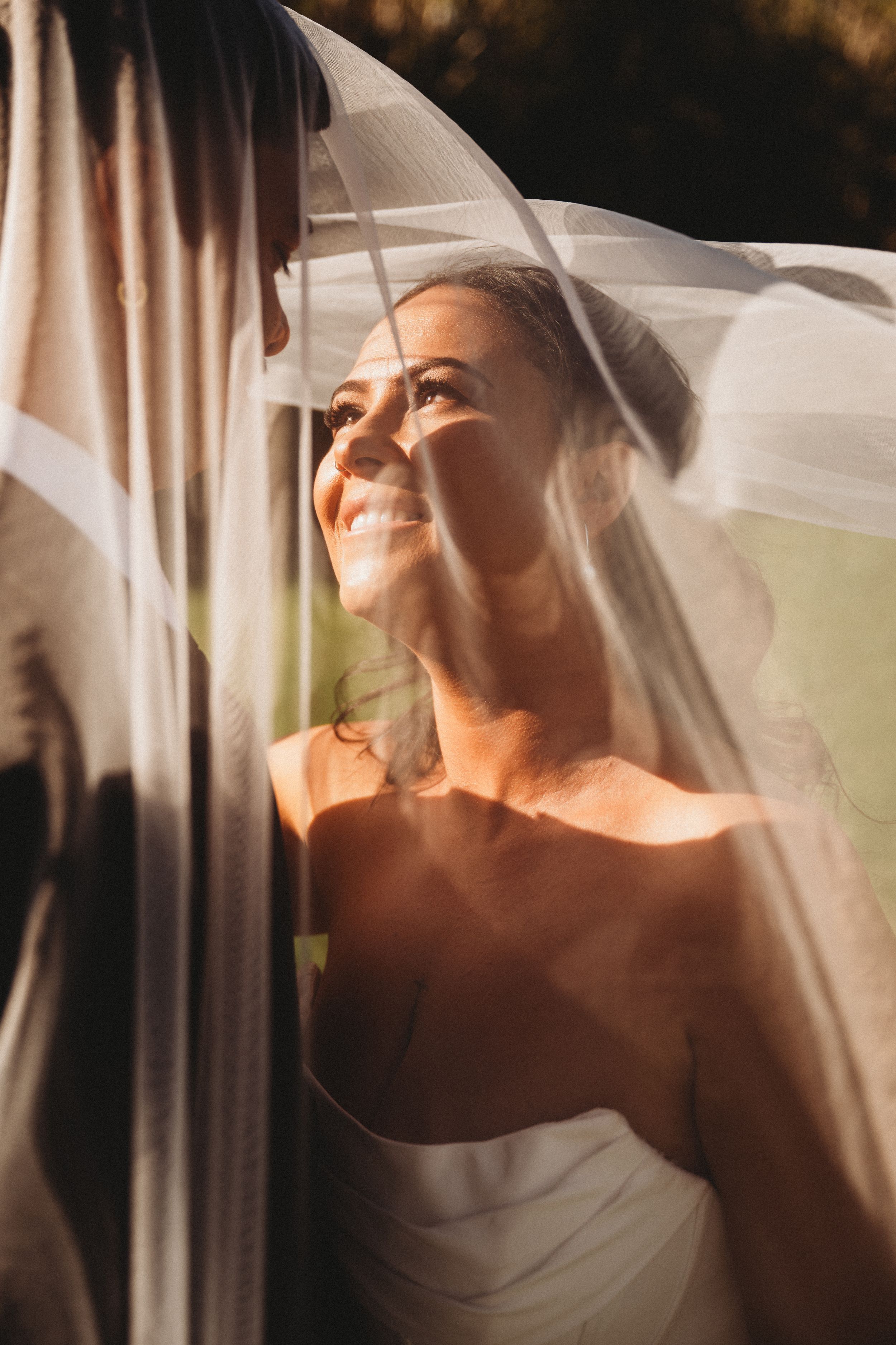 Radiant bride smiling under flowing veil in golden sunlight during outdoor wedding ceremony.