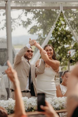 Happy newlyweds raise joined hands at outdoor wedding reception surrounded by white floral arrangements and cheering guests.