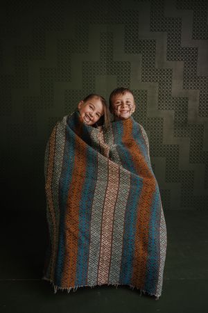 Two smiling children wrapped together in a colorful striped blanket against a dark green patterned wall.